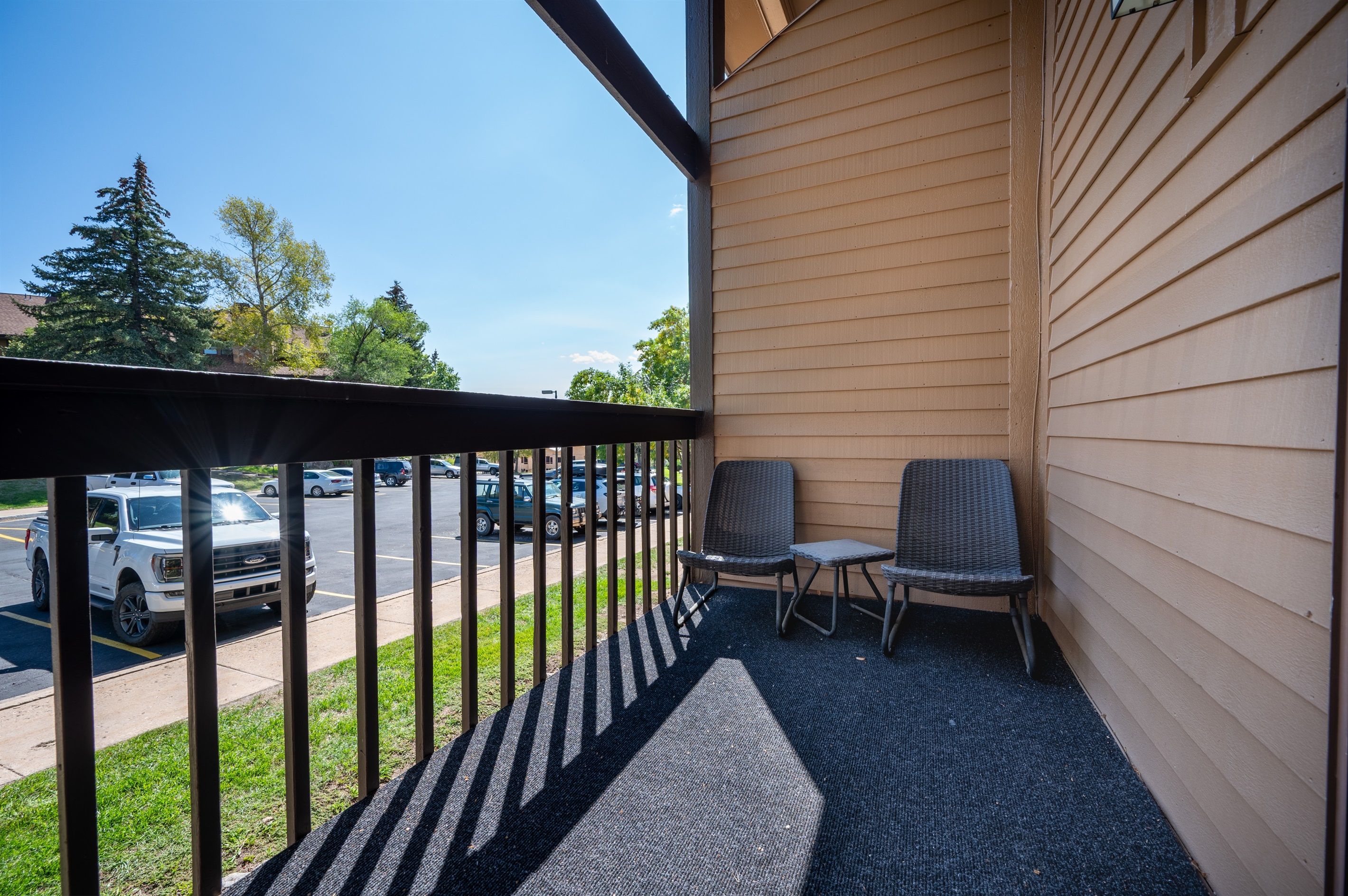 Ein Balkon mit zwei Stühlen und einem kleinen Tisch, mit Blick auf einen Parkplatz und Bäume.