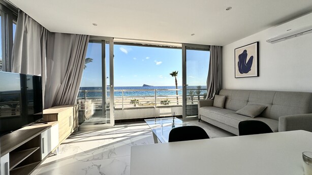 A modern living room with a large TV, a sofa, and a dining table. It features a balcony with a view of the beach and the ocean.