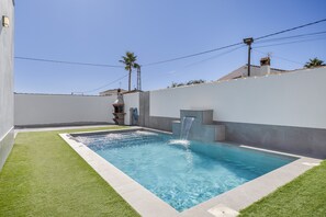 A rectangular swimming pool with a waterfall feature, surrounded by a low wall and a grassy area.