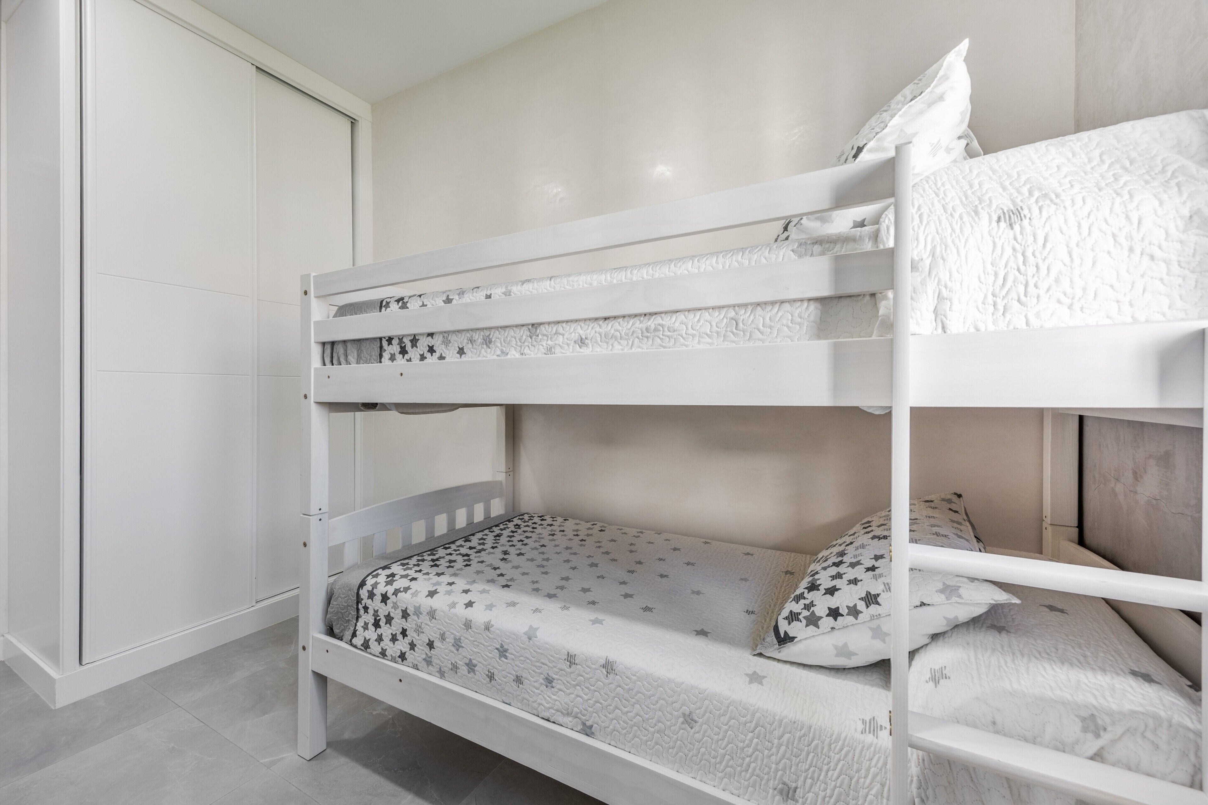 A bunk bed with a star-patterned mattress and pillows, a white wardrobe, and a plain wall.