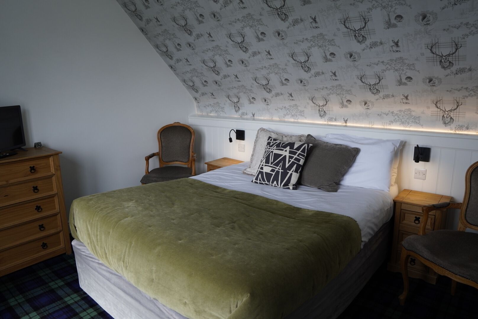 A bedroom with a bed, a dresser, two chairs, a television, and a patterned ceiling.