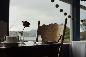 A dining table with a chair, a rose in a glass vase, a menu, and a view of a lake and mountains. - The Lochnell Arms Hotel  (Oban)