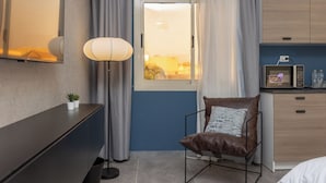 Una habitación de hotel moderna con escritorio, silla, lámpara, vistas al atardecer a través de una ventana y kitchenette.