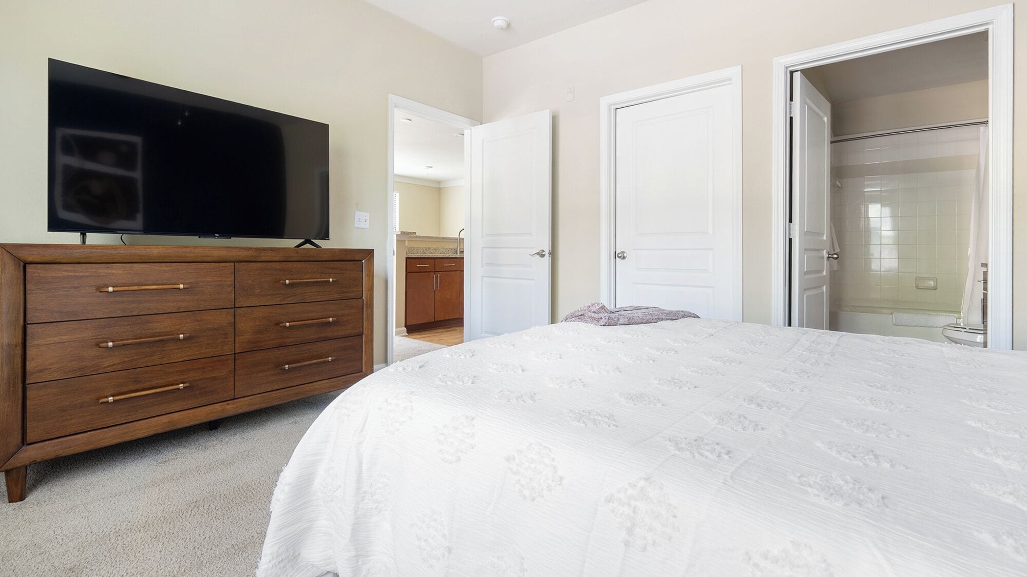 A bedroom with a bed, a dresser, a television, and a bathroom visible through an open door.