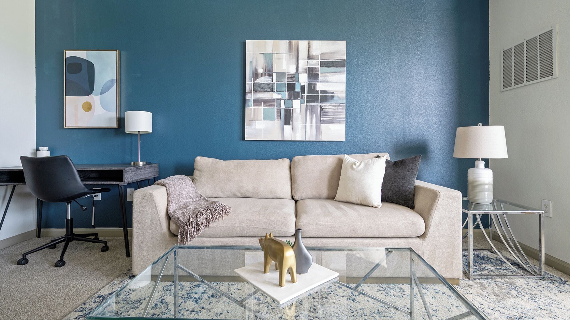 Una sala de estar con una pared azul, un sofá color beige, una mesa de centro de vidrio y un escritorio con una silla.