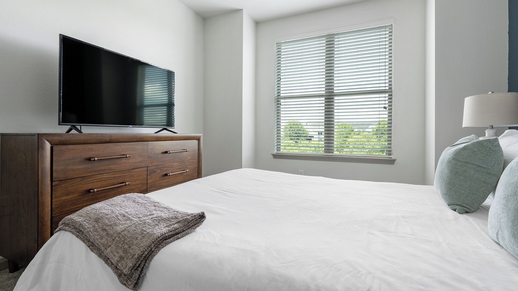 A bedroom with a bed, a TV on a wooden dresser, a window with blinds, and a lamp.