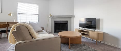 Ein Wohnzimmer mit einer Couch, Couchtisch, Fernseher, Kamin und einem Schreibtisch mit Lampe.