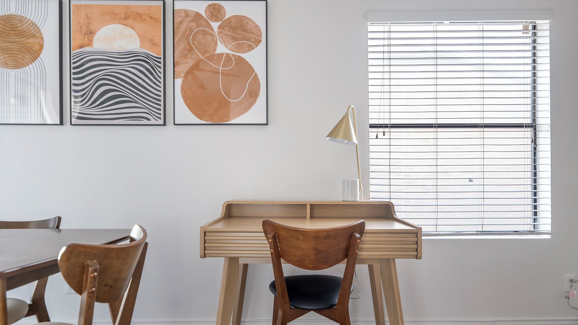 A modern room with a wooden desk, a chair, a dining table, and three abstract paintings on the wall.