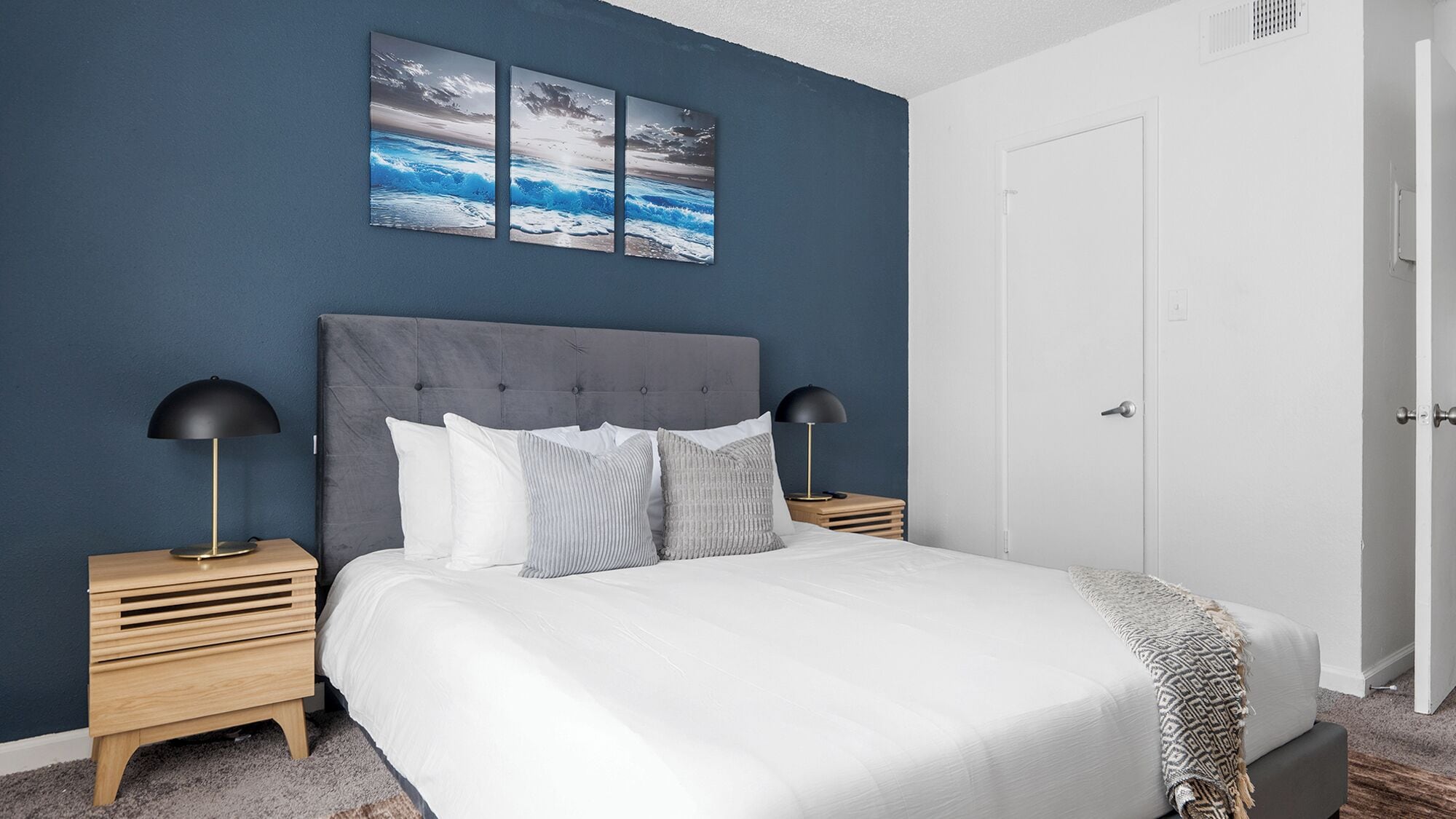 A bedroom with a large bed, two bedside tables with lamps, a dark blue wall, and a door.