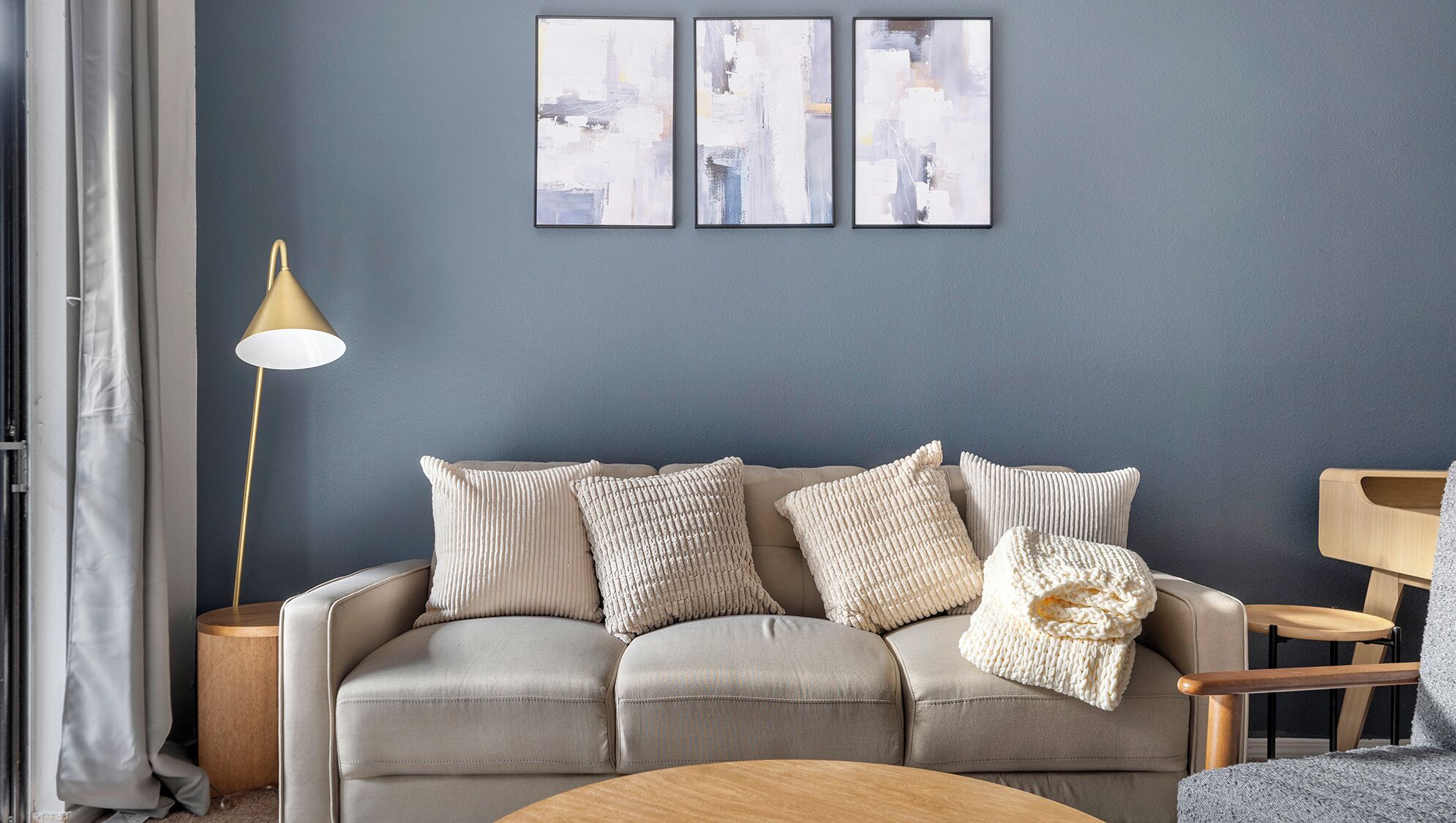 Ein Wohnzimmer mit grauem Sofa, drei Kissen, einem runden Holztisch, einer Stehlampe und drei abstrakten Bildern an der Wand.