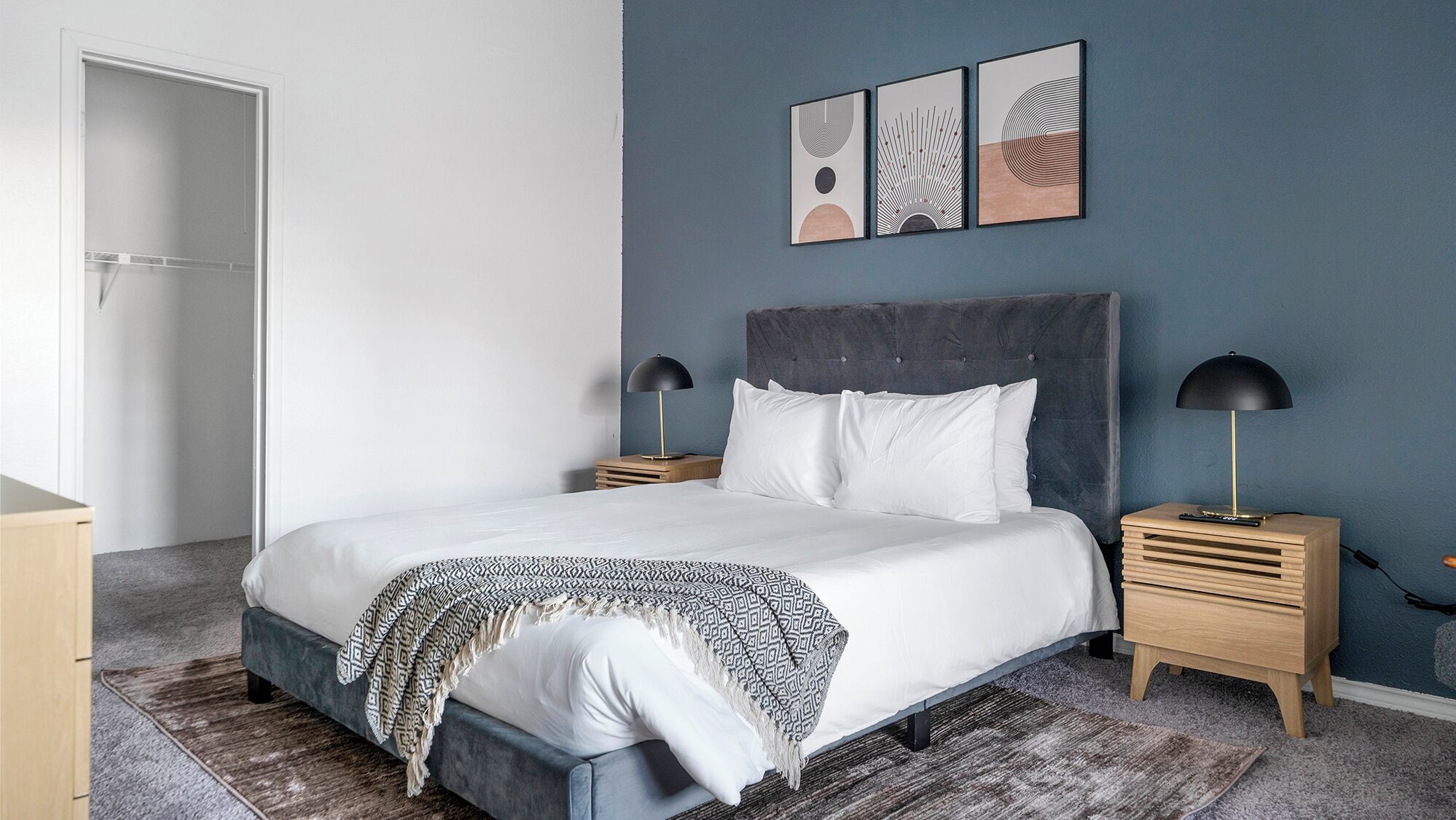 A modern bedroom with a large bed, two bedside tables, a wooden nightstand, and a grey headboard.