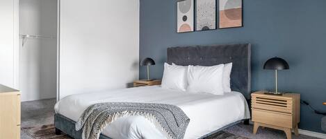 A modern bedroom with a large bed, two bedside tables, a wooden nightstand, and a grey headboard.