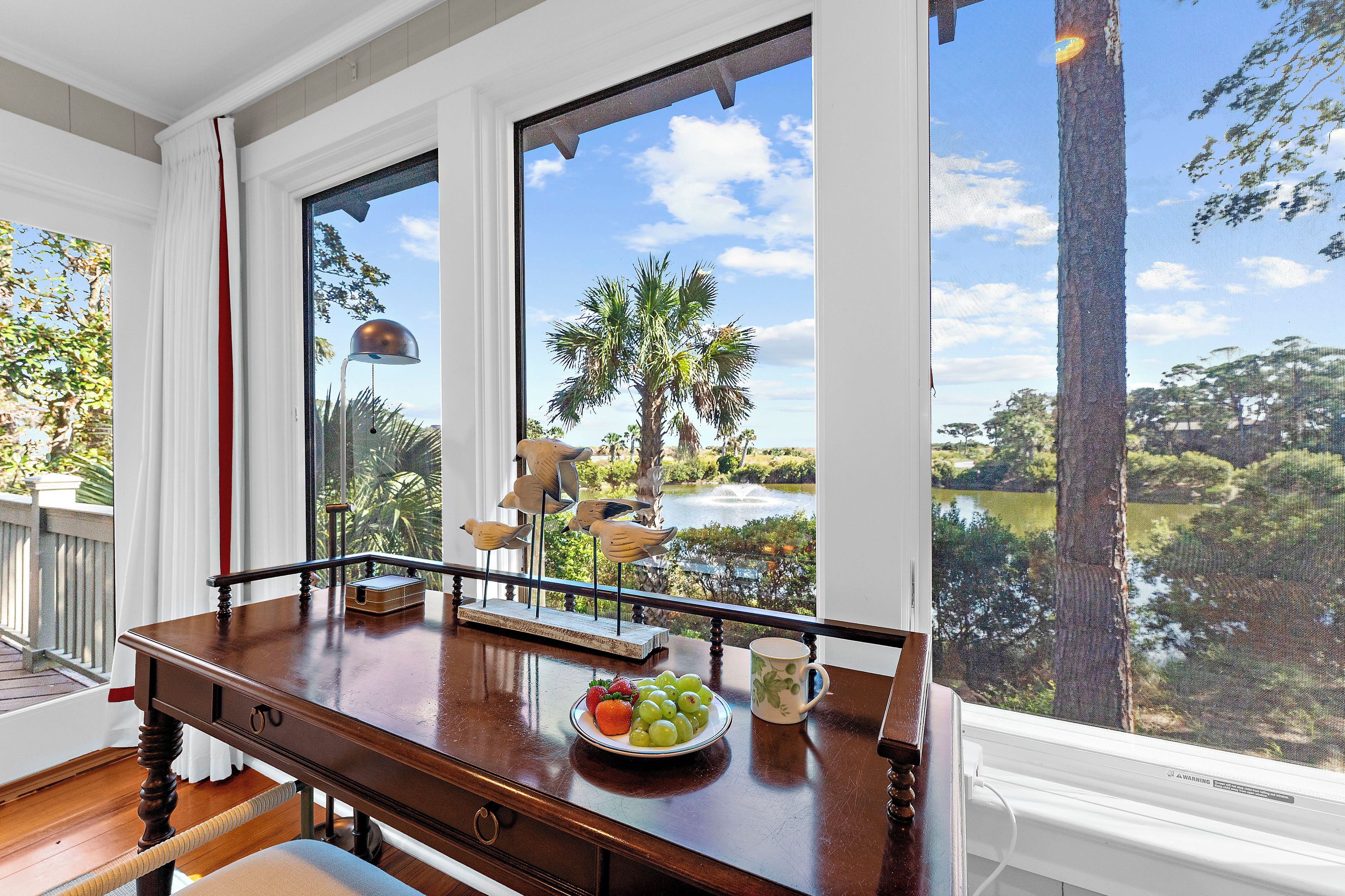 A wooden table with a plate of fruit and a mug, a bird sculpture, and a view of a lake and palm trees through large windows.