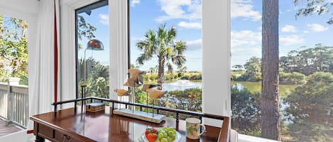 A wooden table with a plate of fruit and a mug, a bird sculpture, and a view of a lake and palm trees through large windows.