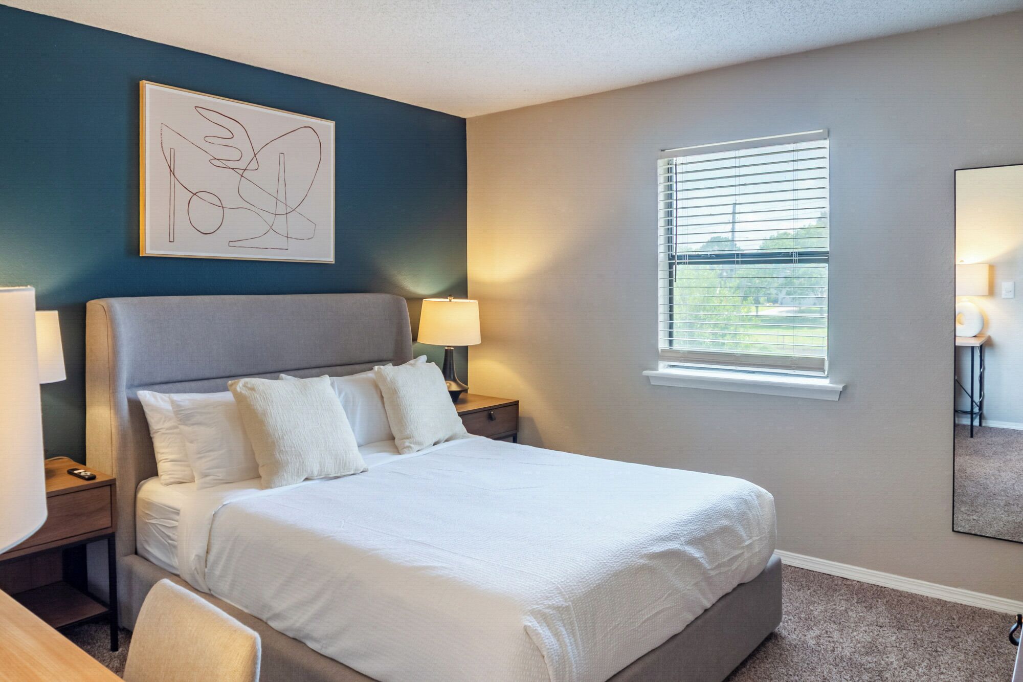 A neatly made bed in a room with a blue accent wall, a window with blinds, and a framed artwork above the bed.