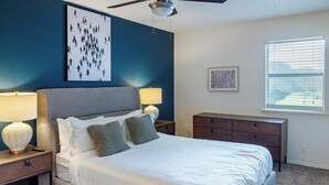 A bedroom with a large bed, two bedside tables with lamps, a dresser, a ceiling fan, a wall art piece, and a window with blinds. - Landing Tulsa near Woodland (Tulsa)