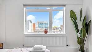 Makuuhuoneessa on suuri ikkuna, patteri, valkoisella peitolla varustettu sänky ja ruukussa oleva kasvi.