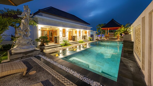 A well-lit swimming pool area with a house in the background, surrounded by plants and a decorative statue. - Cove Dharma Kencana (Kutuh)