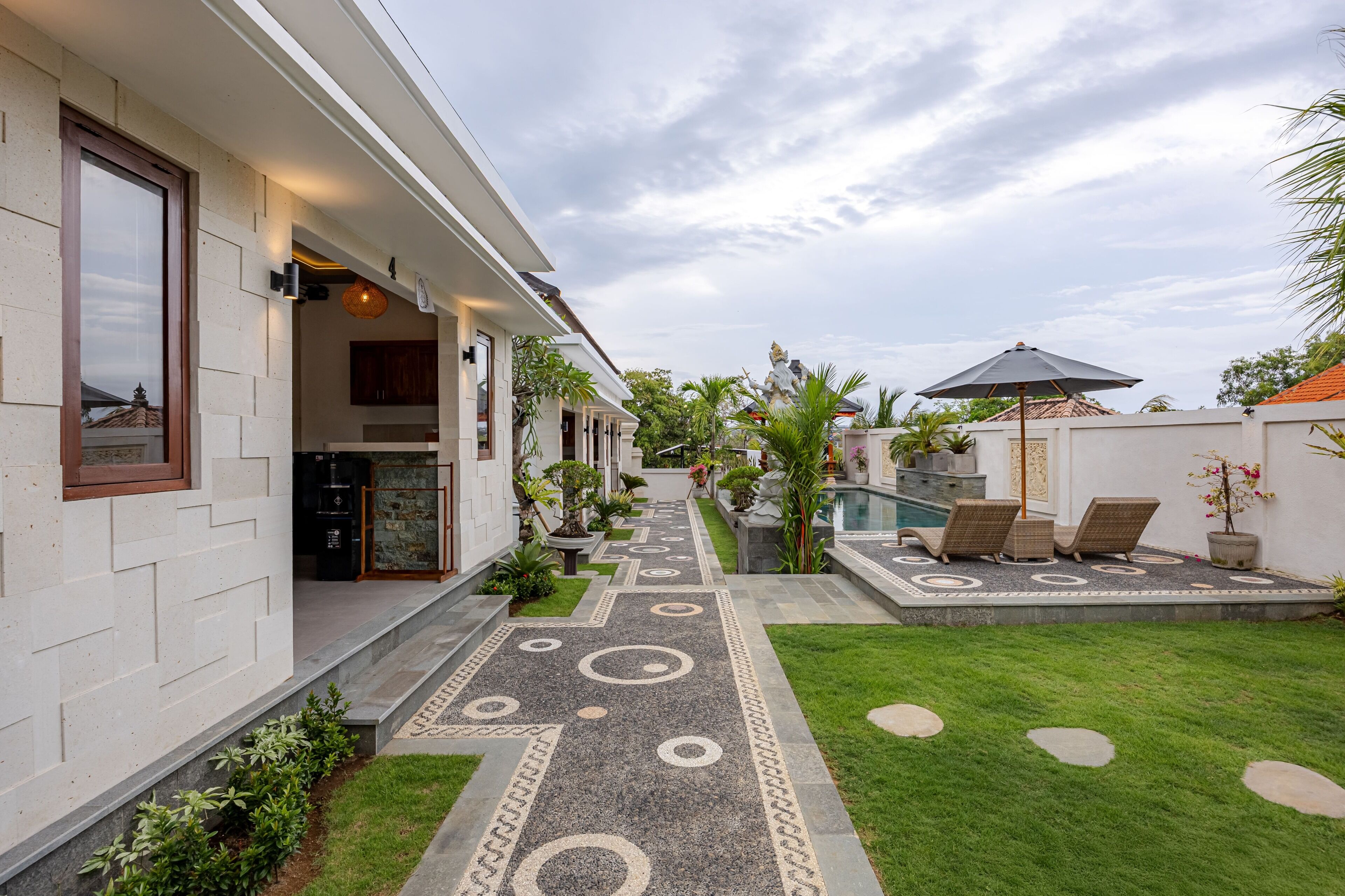 Una casa moderna con un camino pavimentado que conduce a una zona de piscina, rodeada de vegetación y mobiliario de exterior.