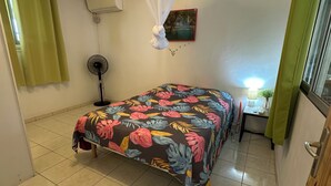 A bedroom with a bed, a standing fan, a mosquito net, a painting, and a window with curtains. - Comfortable Villa with Terrace and Garden – Baie-Mahault Destreland (Baie-Mahault)