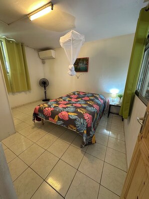 Une chambre à coucher avec un lit, un ventilateur sur pied, une moustiquaire, un tableau et une fenêtre avec des rideaux.