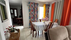 A dining area with a table set for four, surrounded by chairs, a sofa, and a wicker side table with flowers. - Comfortable Villa with Terrace and Garden – Baie-Mahault Destreland (Baie-Mahault)
