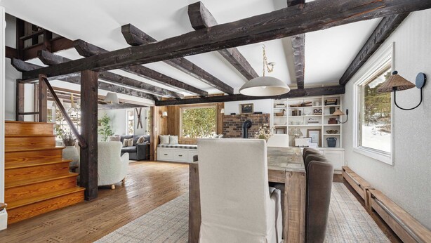 Amplia sala de estar con vigas de madera a la vista, una escalera de madera, una mesa de comedor con sillas y un gran ventanal que proporciona luz natural.