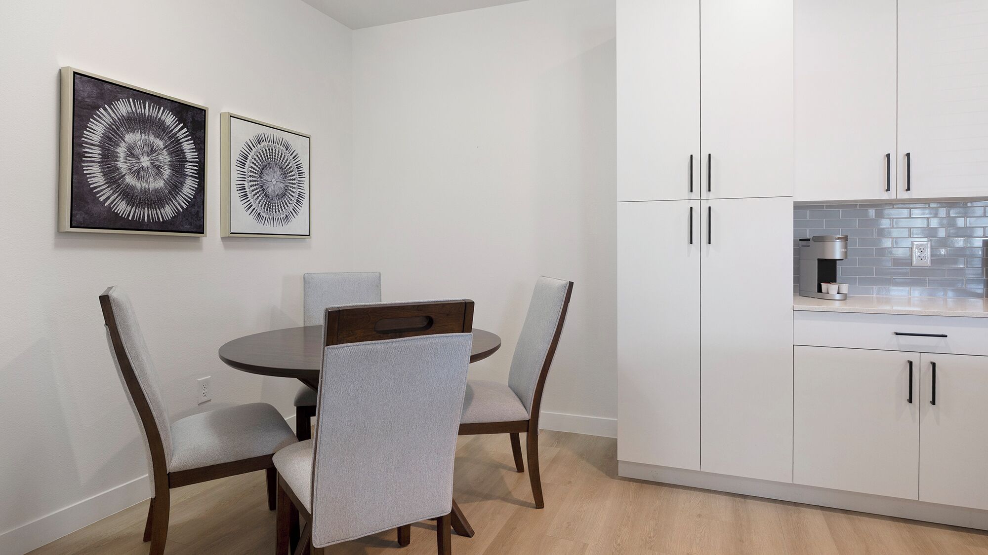 Een moderne keuken met witte kasten, een ronde eettafel en twee stoelen. Er hangen twee abstracte kunstwerken in lijsten aan de muur.