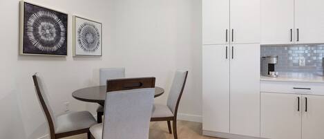 Een moderne keuken met witte kasten, een ronde eettafel en twee stoelen. Er hangen twee abstracte kunstwerken in lijsten aan de muur.