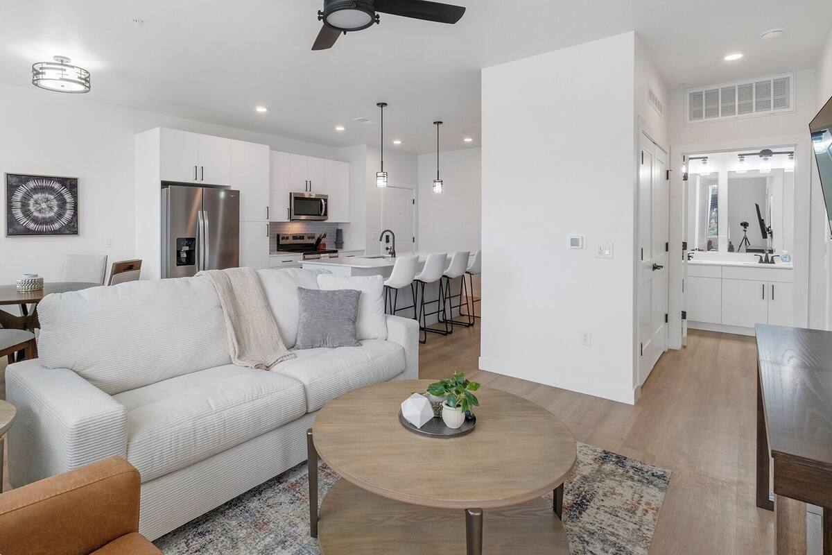 A modern living room with a sofa, coffee table, and chairs. There is a kitchen area with a refrigerator, microwave, and bar stools.