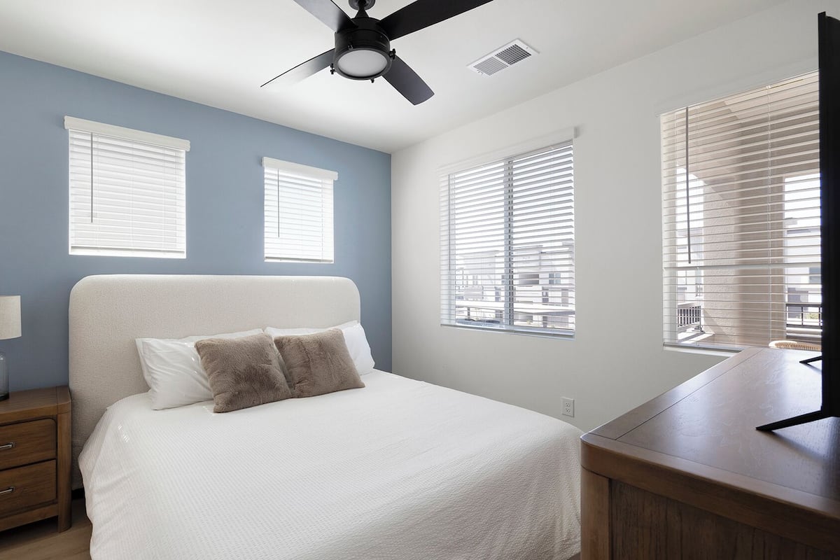 A bedroom with a bed, nightstand, lamp, ceiling fan, and windows.