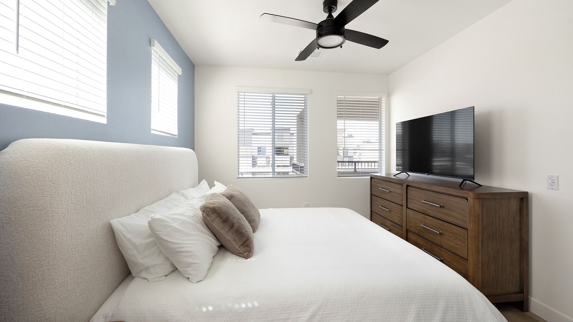 A bedroom with a bed, a TV, a ceiling fan, and windows with blinds.