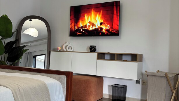 Un dormitorio moderno con cama, espejo grande, chimenea, televisor y mesita de noche.