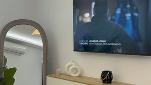 Una sala de estar moderna con un televisor de pantalla plana montado en la pared, una consola blanca, un reposapiés marrón y un espejo grande con marco arqueado.