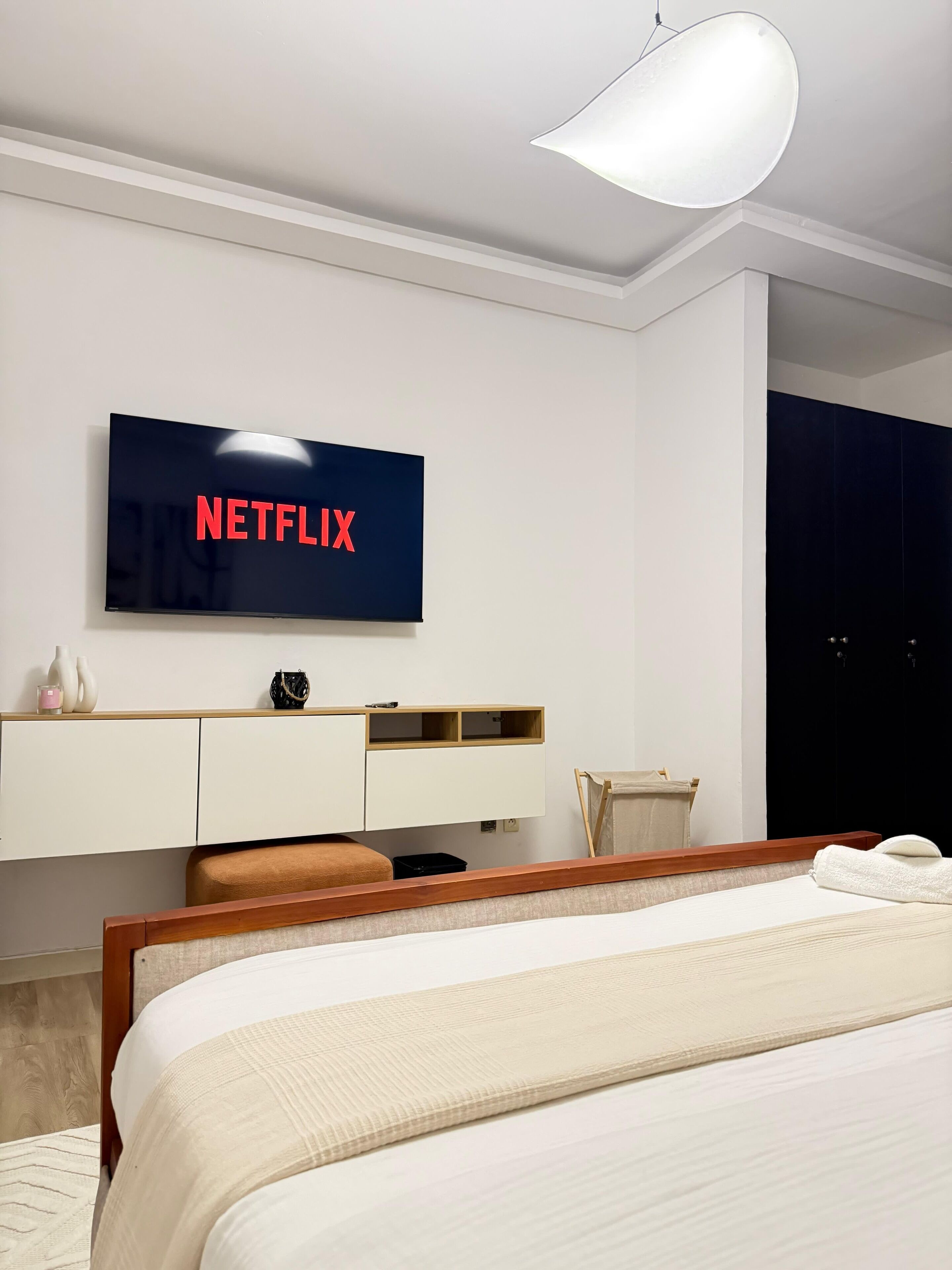 Ein modernes Schlafzimmer mit einem Bett, einem an der Wand befestigten Fernseher, der "NETFLIX" anzeigt, einem Nachttisch mit Lampe und einem bei­ständenden Tisch.