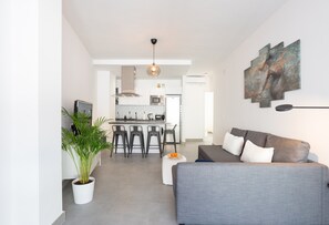 Una sala de estar moderna con un sofá gris, un comedor con taburetes altos y una cocina al fondo.