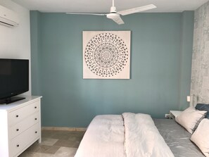 Une chambre à coucher avec un lit, un téléviseur posé sur un meuble, un ventilateur de plafond et une œuvre d’art accrochée au mur.
