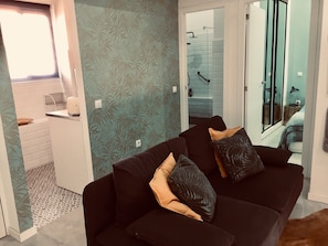 Un salon avec un canapé noir, un papier peint à motifs, une porte de douche en verre et un petit coin cuisine.