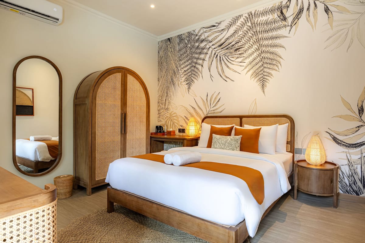 A hotel room with a large bed, a wooden headboard, a wall-mounted mirror, a bedside table with a lamp, and a large fern leaf mural.