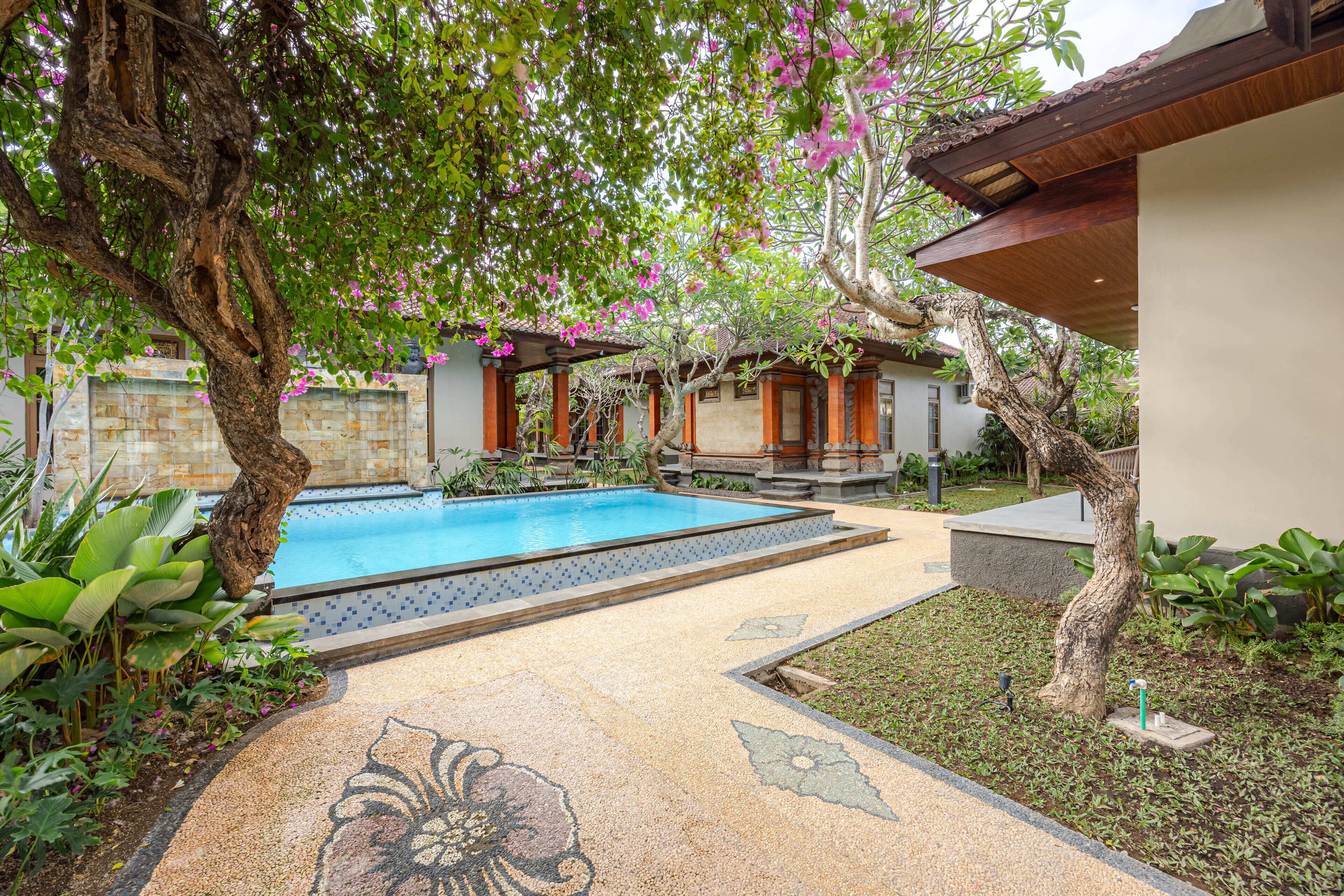 Zona de piscina con suelo de baldosas, rodeada de abundante vegetación y plantas en flor.