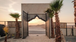 An arched entrance with palm trees, leading to a sandy area with a view of distant hills.