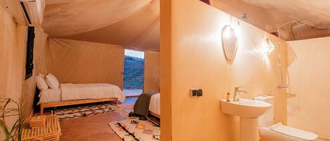 A tented accommodation room with a bed, a bathroom with a sink and toilet, and a view of the outdoors.