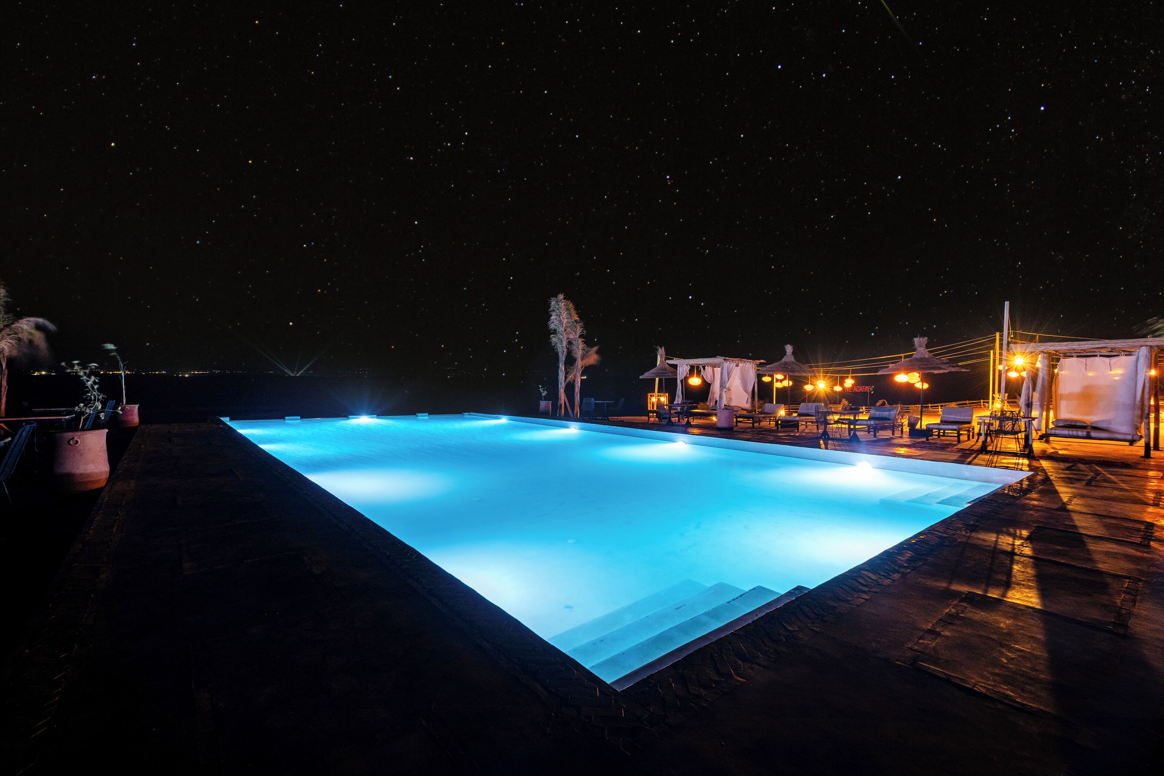 A nighttime swimming pool with a clear view of the sky, surrounded by lounge chairs and a well-lit deck area.