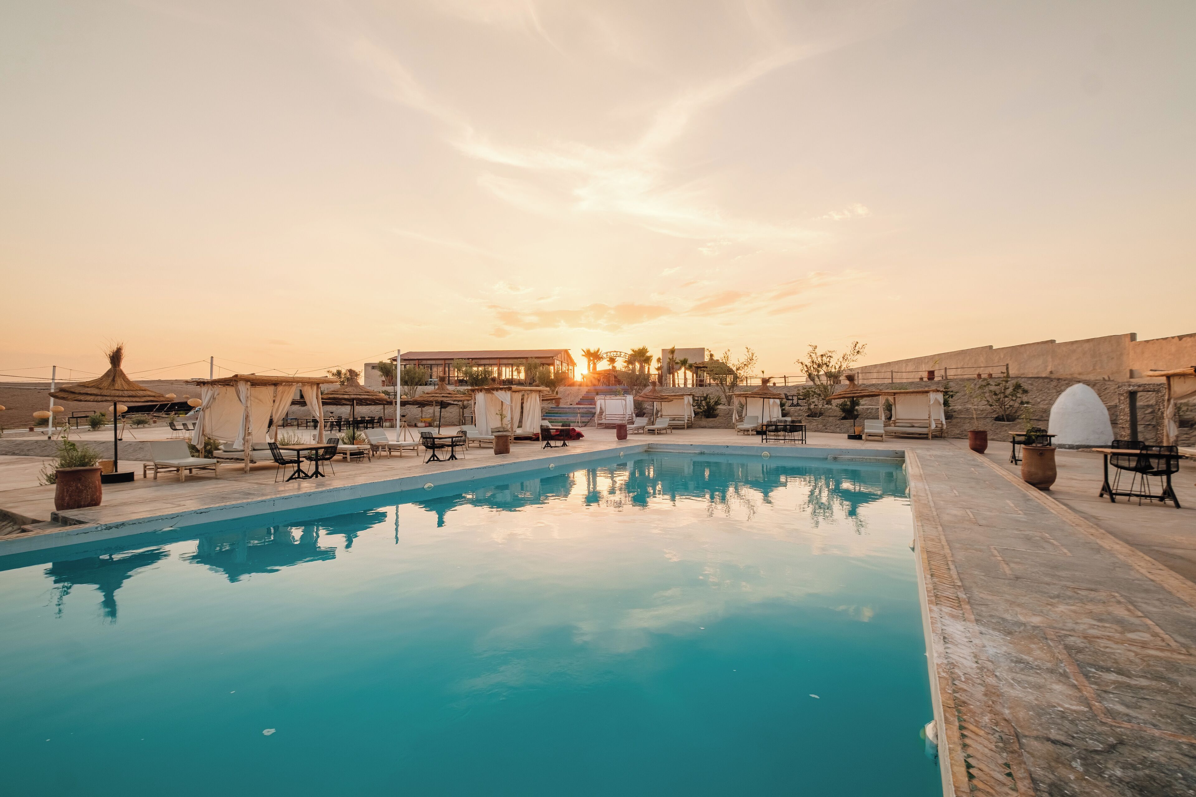 A swimming pool with lounge chairs and umbrellas, surrounded by buildings and a sunset in the background.