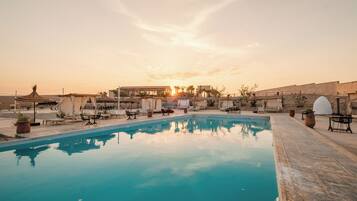A swimming pool with lounge chairs and umbrellas, surrounded by buildings and a sunset in the background.