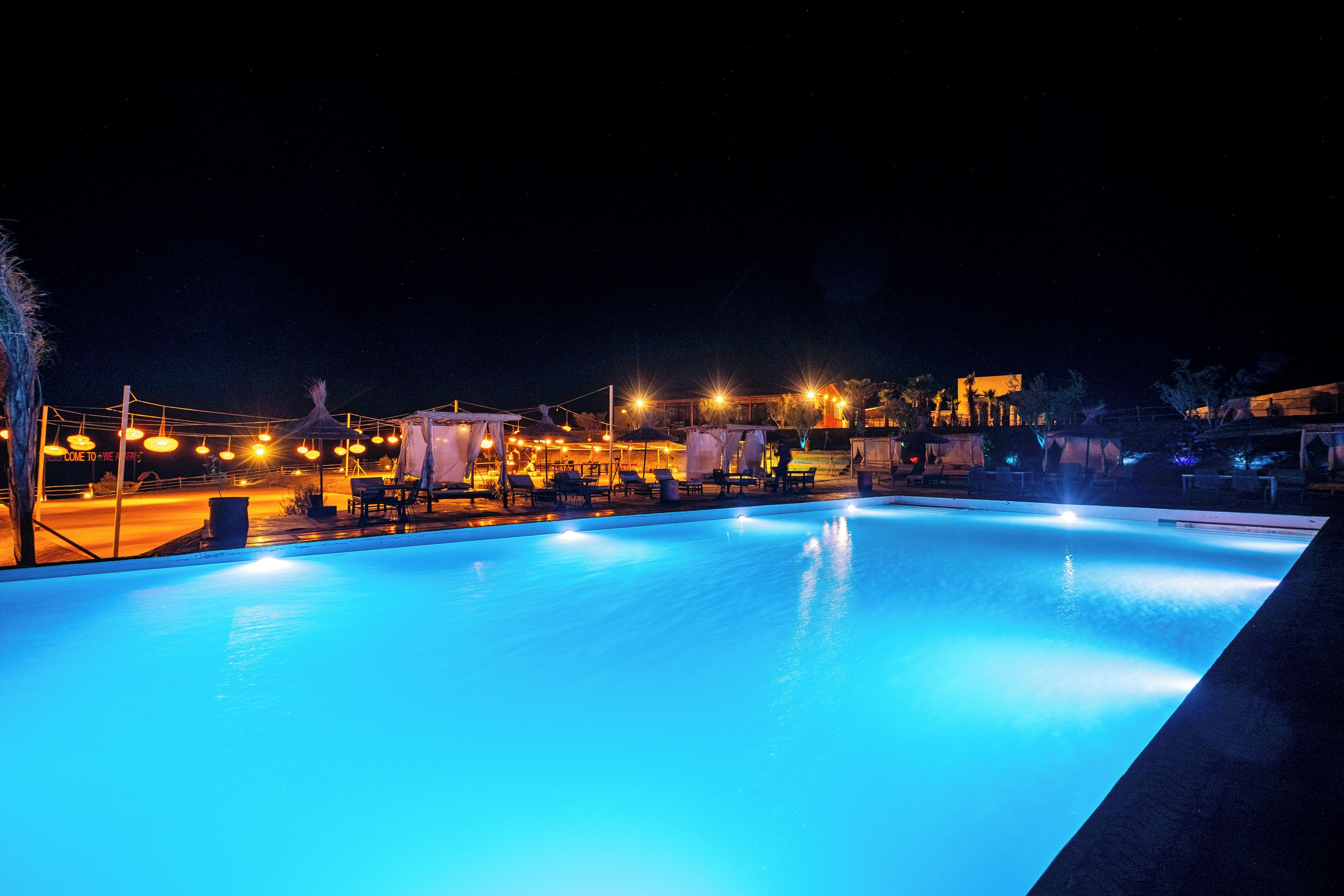 Piscina nocturna con zonas de descanso alrededor y vista a un edificio lejano.