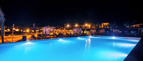 Piscina nocturna con zonas de descanso alrededor y vista a un edificio lejano.