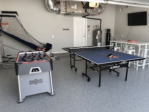 A room with a foosball table, a ping pong table, and a small basketball hoop.