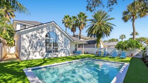 A swimming pool with a white house, palm trees, and blue chairs. - 4BR Pool Game Room | Good Morning Rentals (Jacksonville Beach)