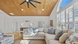 A spacious living room with a high ceiling, a ceiling fan, and large windows with blinds. - 4BR Pool Game Room | Good Morning Rentals (Jacksonville Beach)